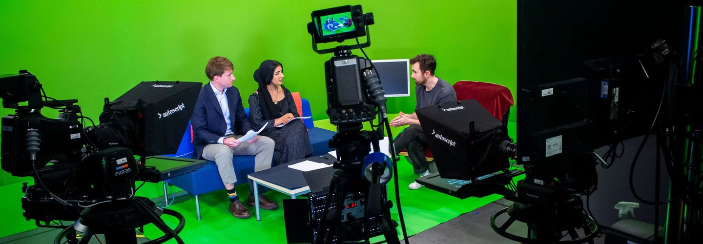 Three people sit on chairs against a green screen, with notes on their laps, appearing to film a broadcast news segment. To the foreground of the image is professional news camera equipment recording
