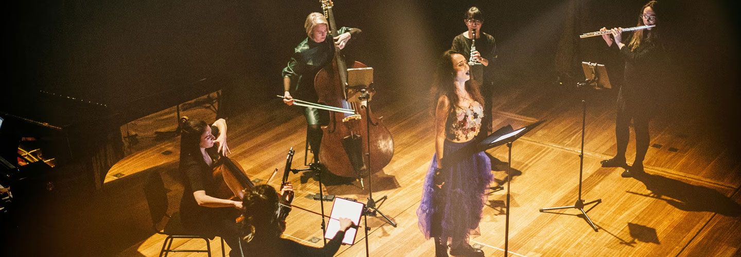 An image of a woman wearing a purple tutu and combat boots and a flowery top singing in front of a stand. All around her are musicians: violonisits, cellists, flautists. They stand on a wooden stage which is dark in every corner