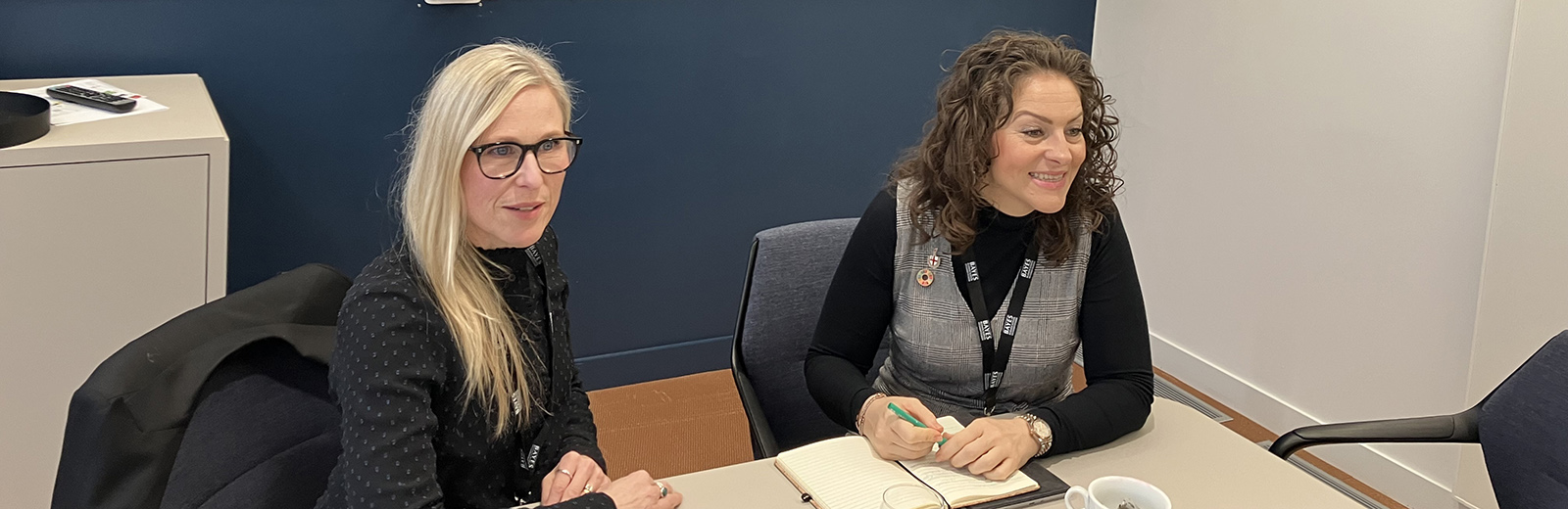 Two women are sitting talking with a screen behind them
