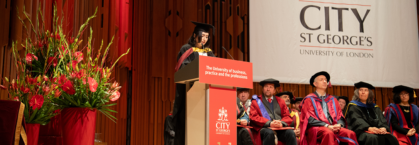 A woman stands at a podium on stage dressed in cap and gown with academics sat nearby on stage