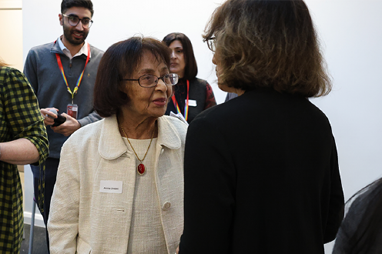 A woman in a suit talks to another person. There are people in the background. 