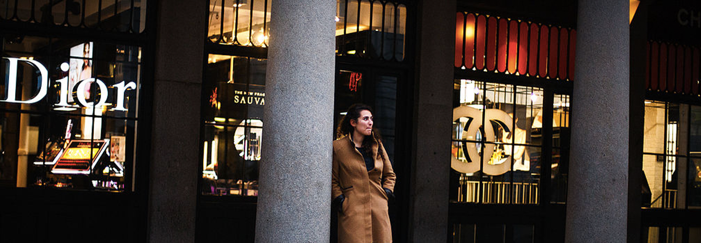 A woman leans against a pillar in a busy brightly lit street at night in front of the Dior shop window