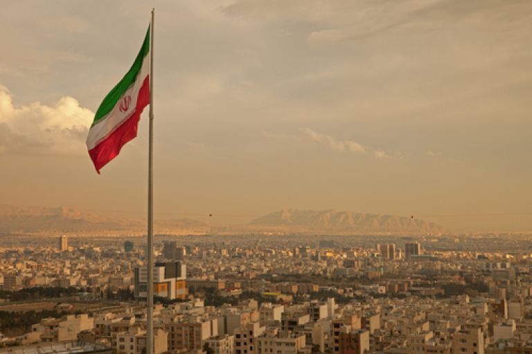 Iran flag waving in the wind above skyline of Tehran lit by orange glow of sunset.
