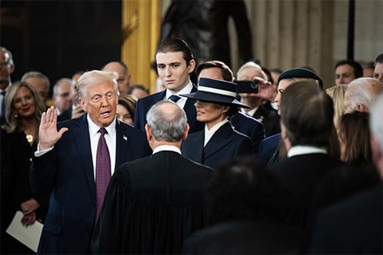 An image of Trump at his inauguration. He holds his hand up and has his mouth open as he takes the oath. Around him in smart clothing is his family -- in particular his son Baron who towers over everyone, and his wife Melania, who smiles and wears a large hat that covers her eyes. We can see the top of his daughter Ivanka's head. Towards the right and partly covered up are former President Biden and former VP Harris. 