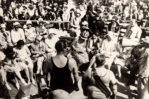 A black and white photo showing two men in bathing costumes being looked at by several dozen women sitting down