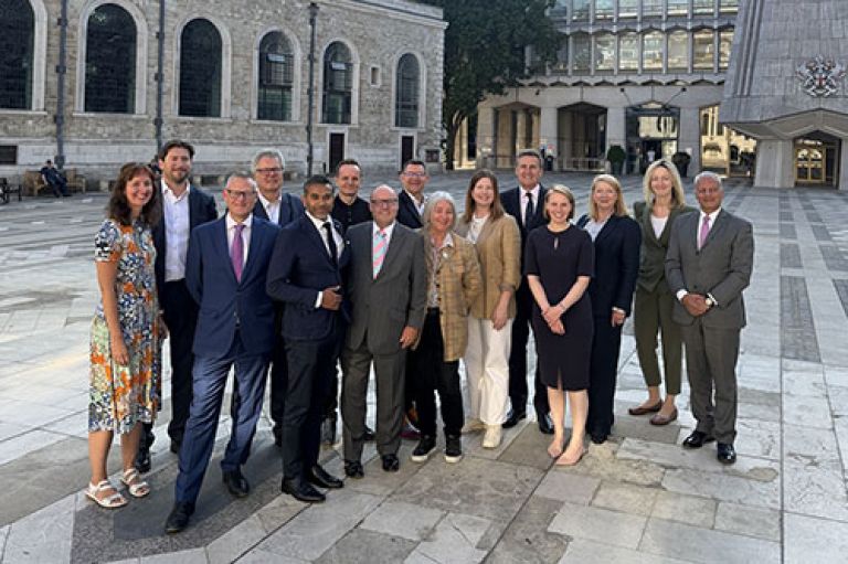Members of the City of London Corporation's Destination Advisory Board stand in the middle of Guildhall Yard and smile to camera. Behind them is the  St Lawrence Jewry church, which has ancient stones and a tower with a clock with golden hands and numbers. To one side is a more modern building made of concrete and glass, with the crest of the City of London on its front. The group wears smart dresses and suits.