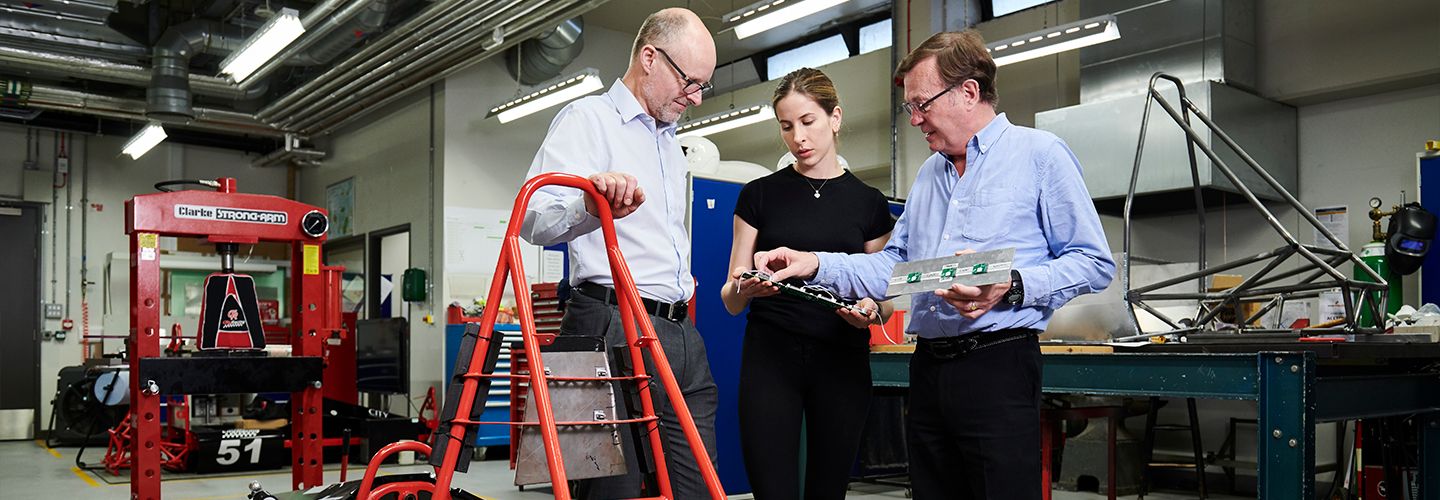 Research student and professor's holding electrical device beside racing vehicle