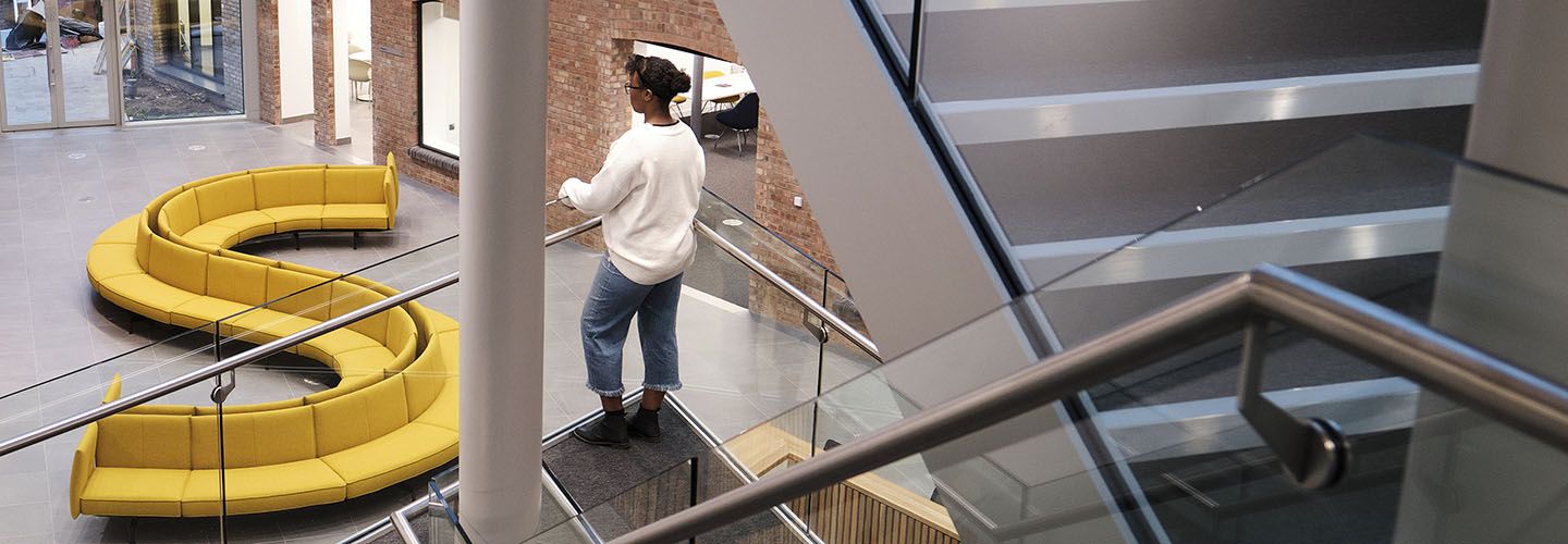 Internal of the Law School Building looking down from mezzanine at yellow sofas and stairs with a person looking over the balustrade on the left.