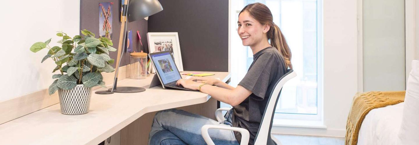 Young female student sat at desk with laptop in single room at Eleanor Rosa House.