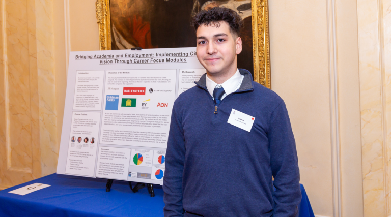 Leo Salem wears a blue jumper, a blue tie and a white shirt. He stands in front of his poster, which is large and covered in text and logos, propped up on a blue table. Behind the poster is a painting with a gold frame, against a gold wall. 