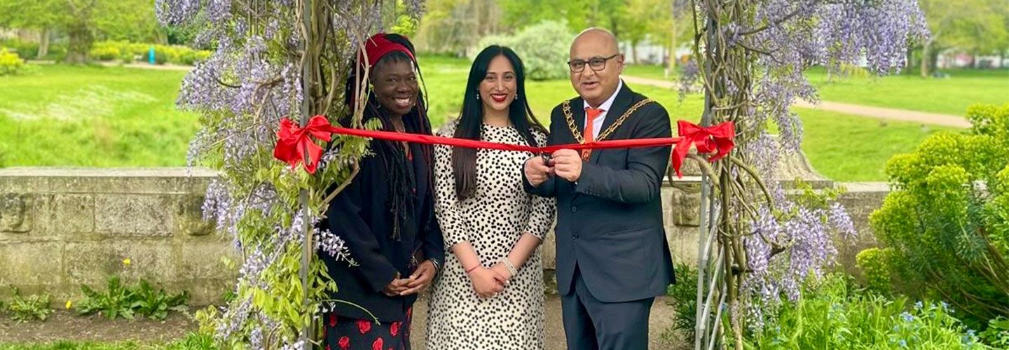 Three people stand under a wooden arch with wisteria growing on it. At their shoulders is a red sash. The Mayor of Ealing holds scissors to cut this. They stand in a green park.