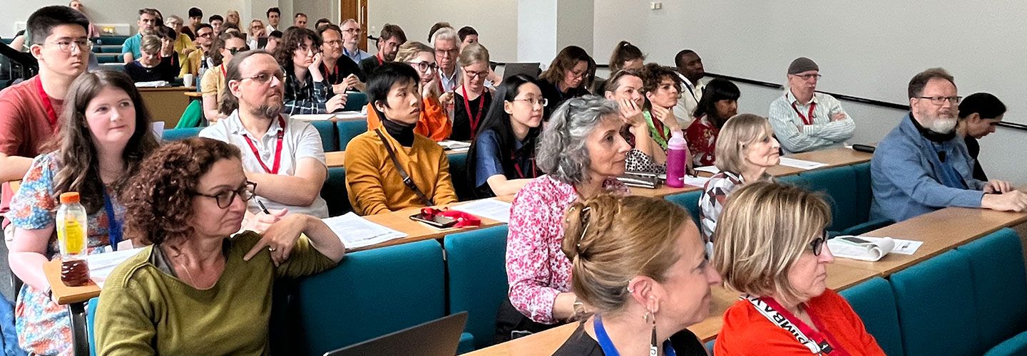 A diverse and full room of attendees sit in rows and look off camera towards the speakers