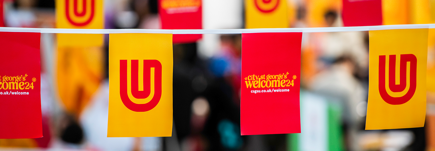 Red and yellow banners hang across a large hall and read 'City St George's Welcome'