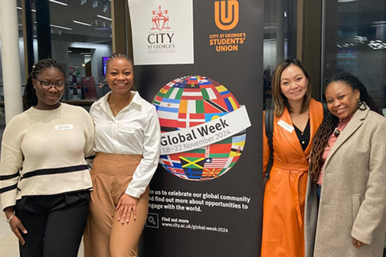 A group of women smile together near a Global Week banner. 