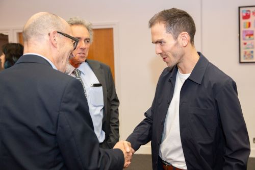 Three men in a crowded room - two are shaking hands