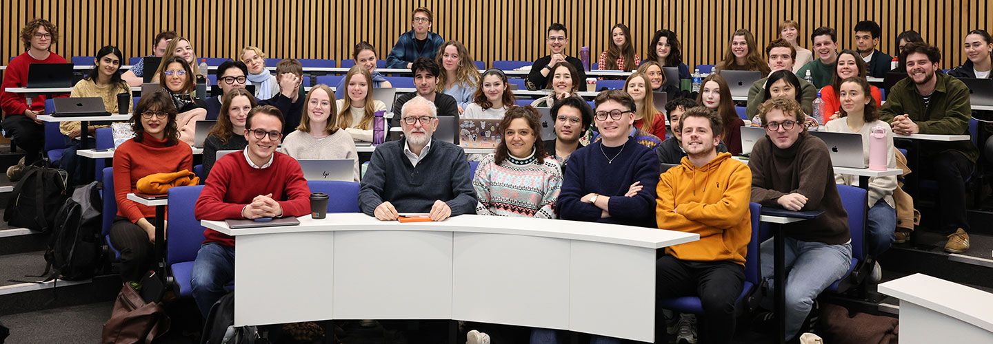 Jeremy Corbyn MP sits among a group of students. The students are on rows of blue seats in front of white desks against wooden walls