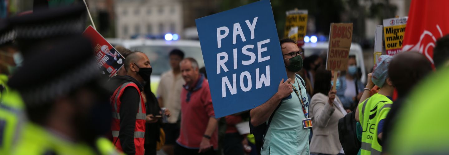 An image of National Health Service (NHS) workers stage a protest demanding pay raise outside Downing Street in 2020. One person holds a sign which reads 