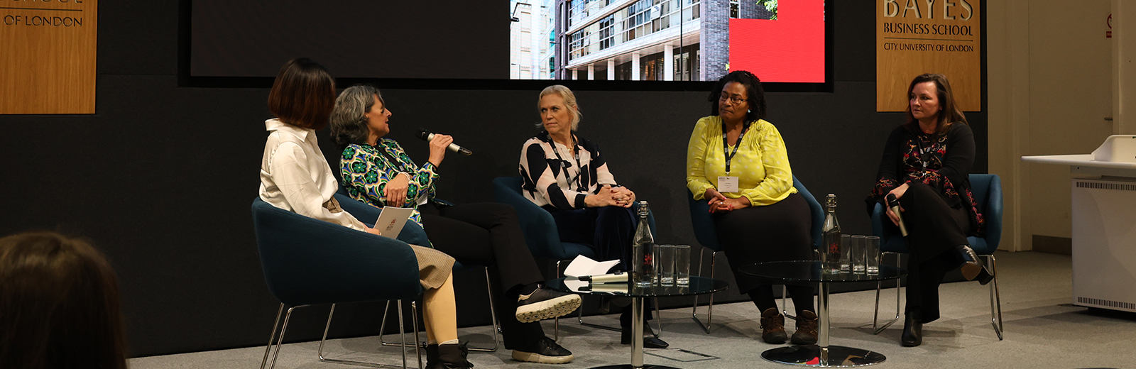 Five women, one of whom has a microphone, sit on a stage 