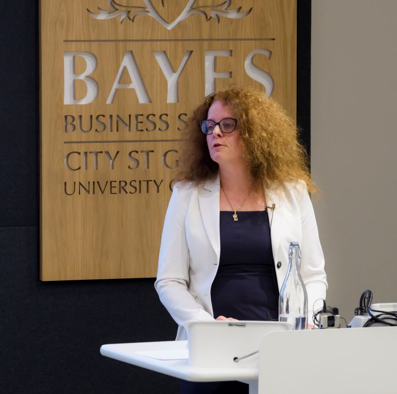 Professor Schnabel delivers her lecture at a podium in front of a Bayes Business School sign