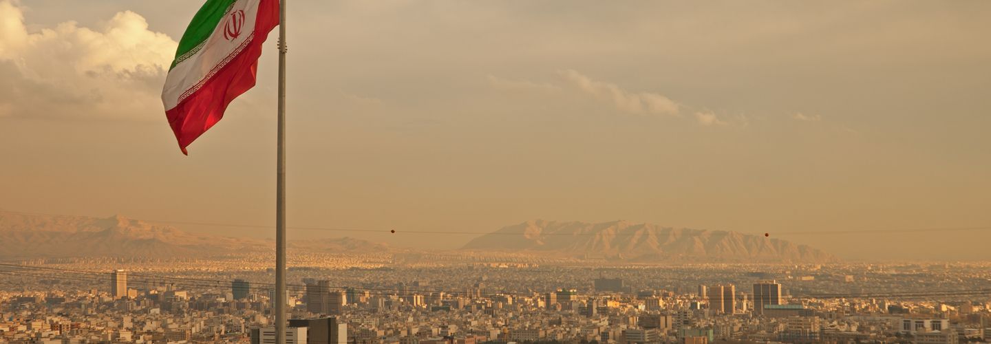 Iran flag waving in the wind above skyline of Tehran lit by orange glow of sunset.