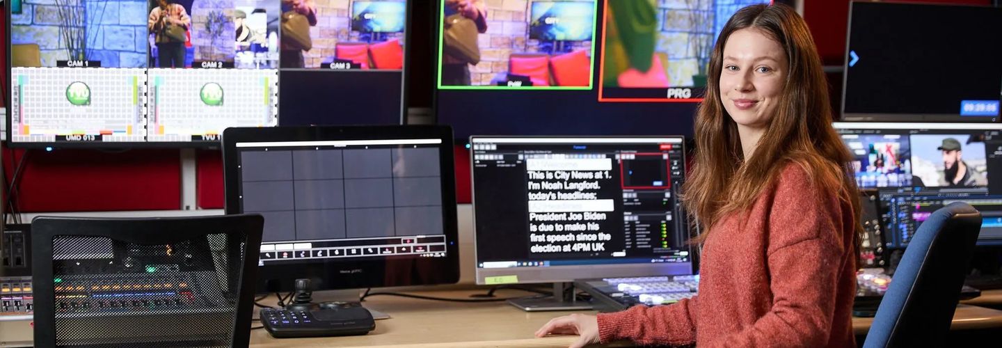 Young woman sitting in front of a bank of screens, on of which displays a newsreader's prompt.