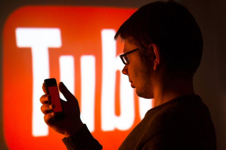 A man wearing glasses holds a mobile phone in his hand. Behind him in red letters is the word 'Tube' from the logo YouTube