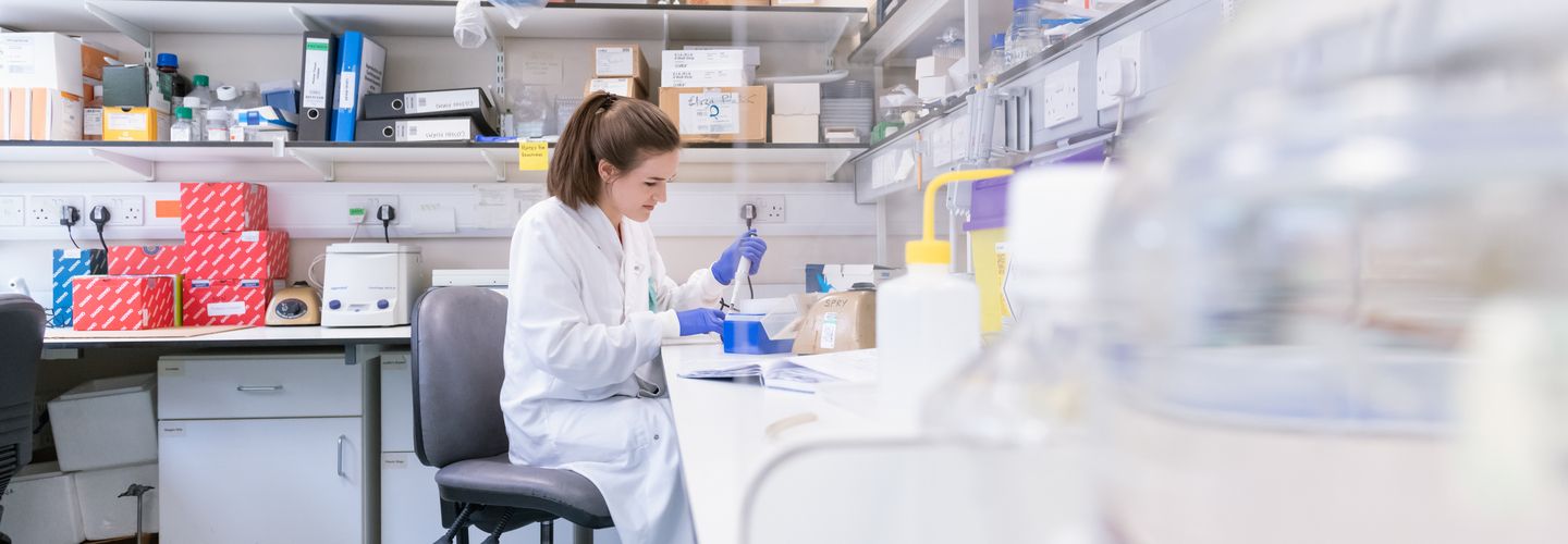A woman in a lab coat and gloves works intently at a lab bench, surrounded by scientific equipment and shelves of supplies, conveying focus and precision.