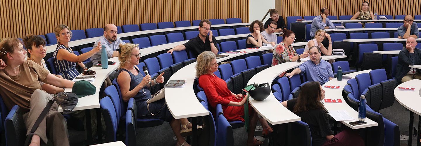 The audience looks at Professor Jane Martinson, who holds the microphone to ask a question. They are all sitting on rows of seats in a horseshoe shaped lecture room.