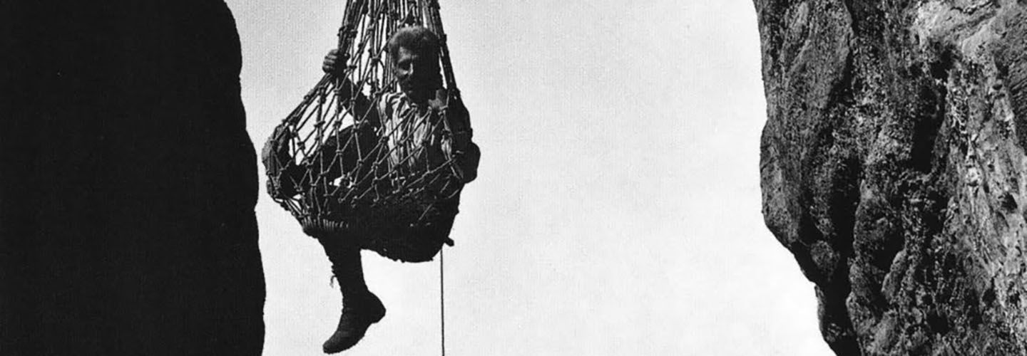 A man sitting in a large net suspended between two cliffs. Black and white photo.