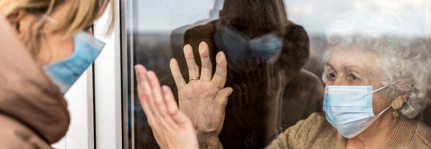 Elderly lady with a surgical mask on looking at a carer with a mask on standing outside her window.