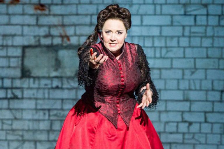 Image of Susan Bickley performing. Her hair is piled high and also goes down to her shoulders. She is wearing a full red dress with a darker red corset on top, with long black sleeves. Her hand is stretched out towards the camera.