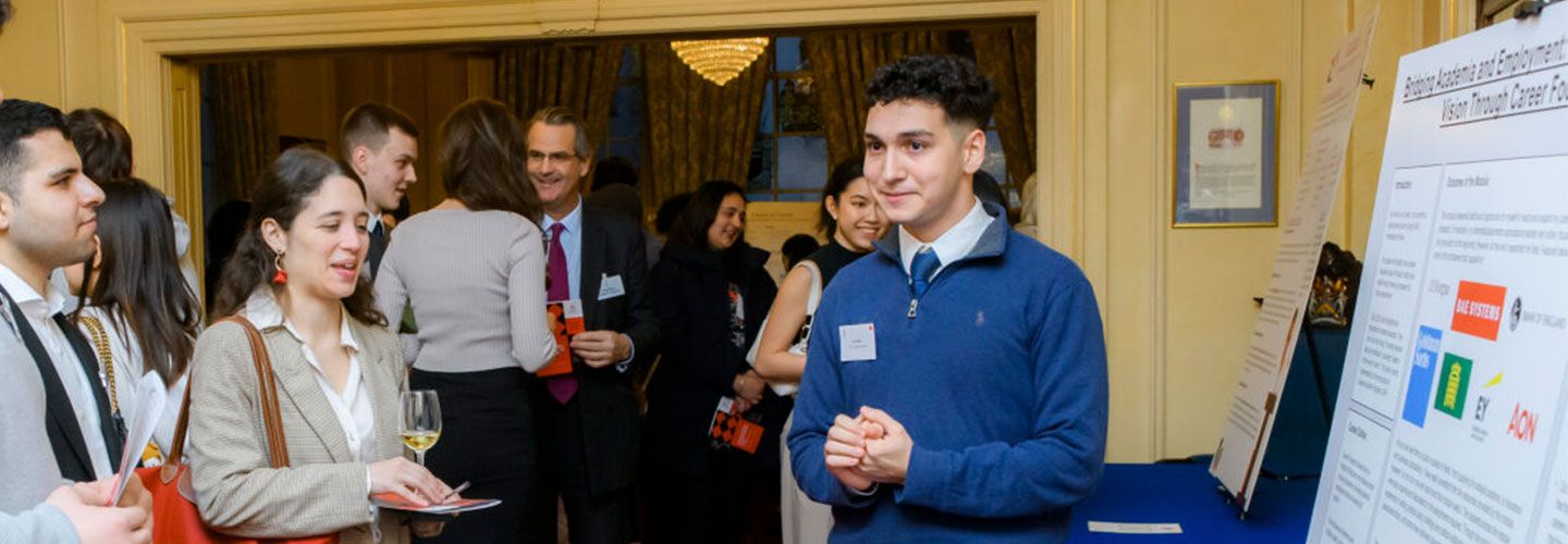 Leo Salem wears a blue jumper, a blue tie and a white shirt. He stands in front of his poster, which is large and covered in text and logos, propped up on a blue table. He is presenting his paper to a crowd of smartly dressed students. Behind him, more people mingle and talk. The walls in the room are gold