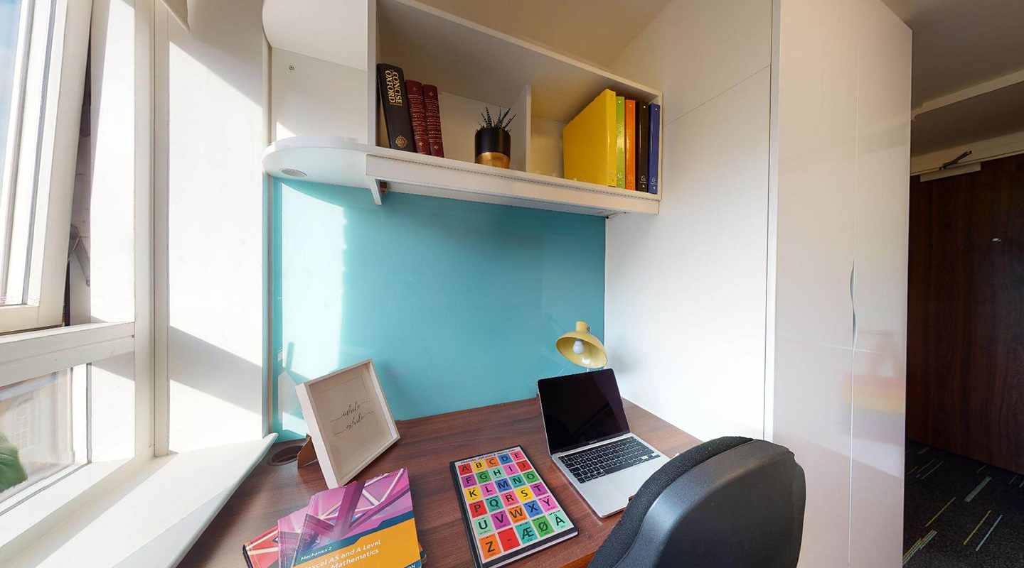 View of the desk and desk chair next to a window, with overhead shelf, lamp, and a wardrobe to the side.