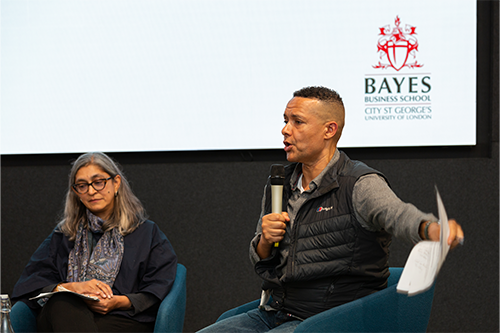 Clive Lewis with a microphone while Dr Sujata Visaria listens in front of a screen with Bayes Business School on it