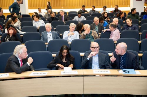 Three men and a woman sit in the front row of an auditorium chatting with a number of the audience behind them