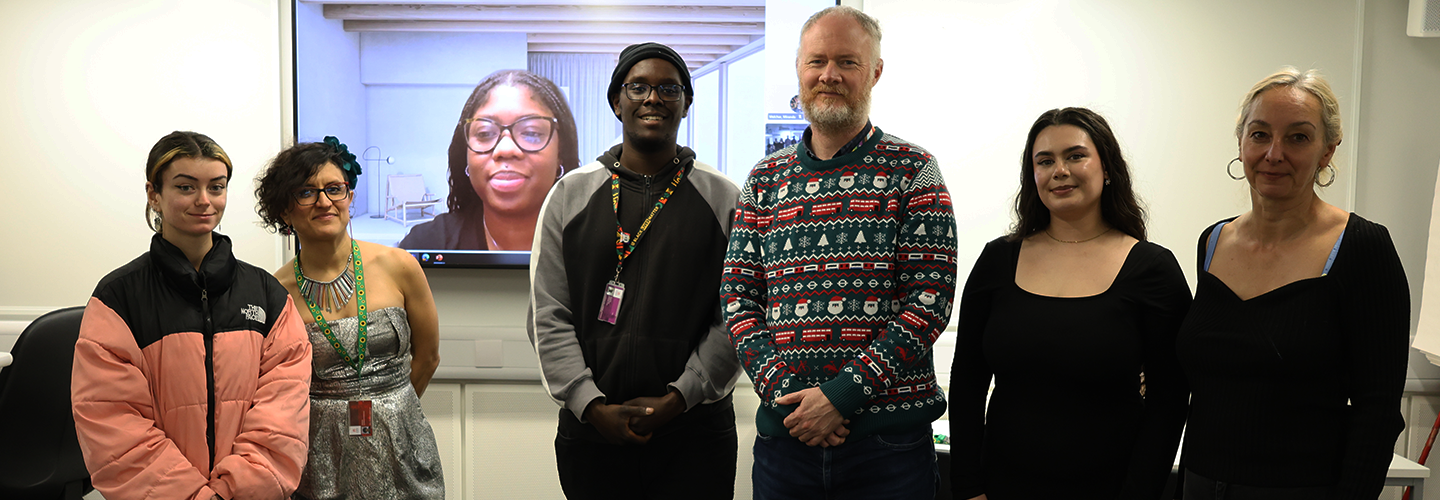 A group of staff and students stand together, with one member of staff appearing on a TV screen behind them, at a hybrid event