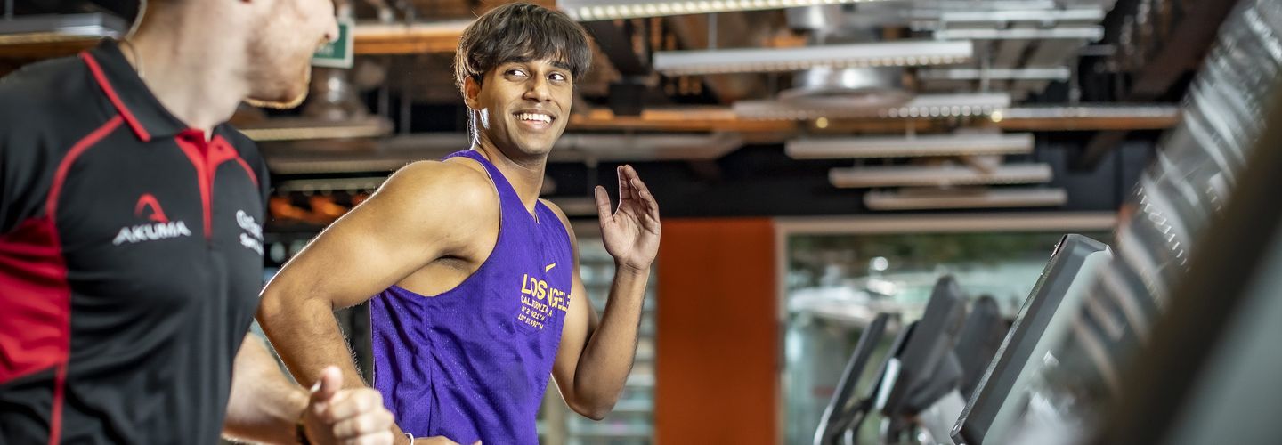 A man smiles while on a running machine, another man is wearing a gym work outfit and appears to be his personal trainer. 