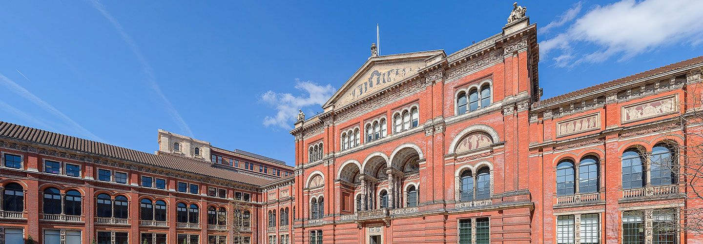 An image of the V&A Museum Central Garden. It is a red building with arches and Greek-resembling drawings of various figures below the arches. Towards the bottom of the image is a flat fountain. The sky is bright blue.
