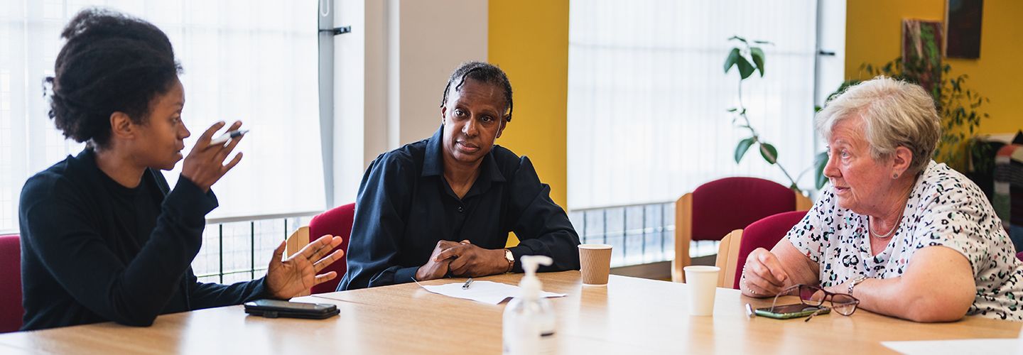 Two Black women and an elderly white woman in discussion around a table, with notes and smartphones to hand.