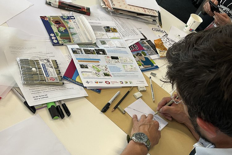A man drawing at a table cluttered with craft materials.