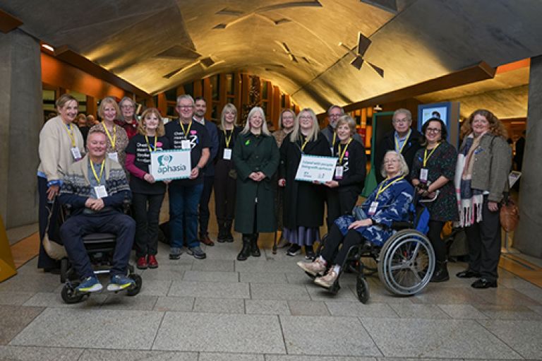 A group of people standing or in wheelchairs facing the camera. The group are arranged in the hall within the Scottish Parliament building in Edinburgh. One person holds a sign showing the word 'aphasia'. Another holds a sign showing 'Chest Heart and Stroke Scotland'