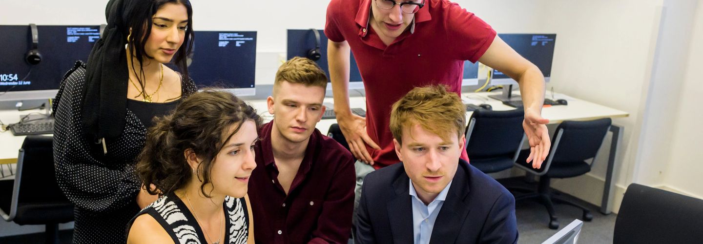 A group of five young adults gathered around a computer in a classroom or computer lab, focused on the screen.