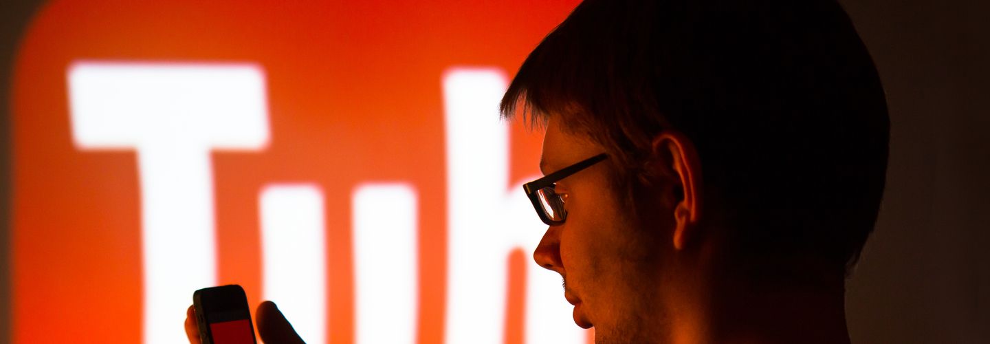 A man wearing glasses holds a mobile phone in his hand. Behind him in red letters is the word 