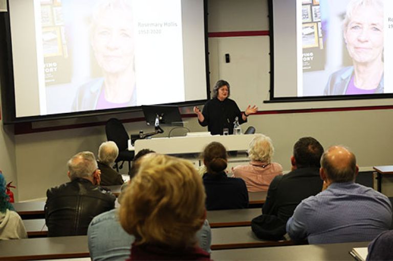 An image of Prof Wendy Pullan standing at with her hands out to either side. Behind her, two digital screens display a picture of the late Prof Rosemary Hollis. In front of Wendy, an audience sits in rows in an amphitheatre-style lecture room. 