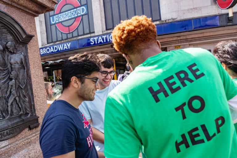 Man in green shirt “HERE TO HELP” converses outside Tooting Broadway subway station entrance.