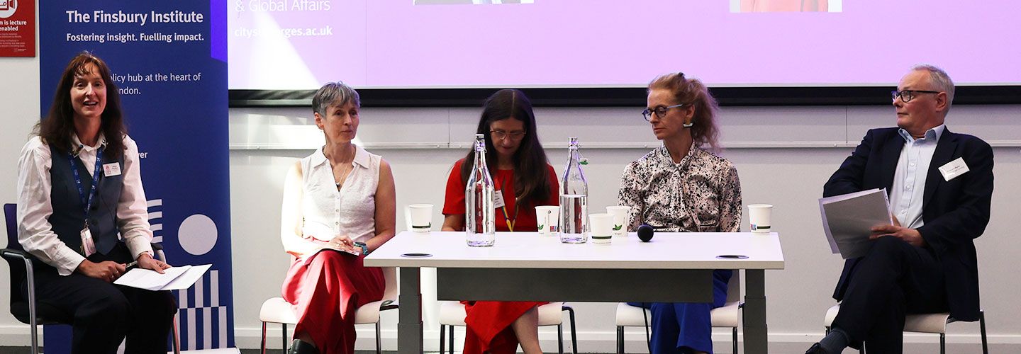 Professor Diana Beech (left) speaks to an audience. Behind her is a banner with the logo of the Finsbury Institute. To her right, sitting in a row, with a table in front of them, are university leaders. Prof Beech is moderating a panel event