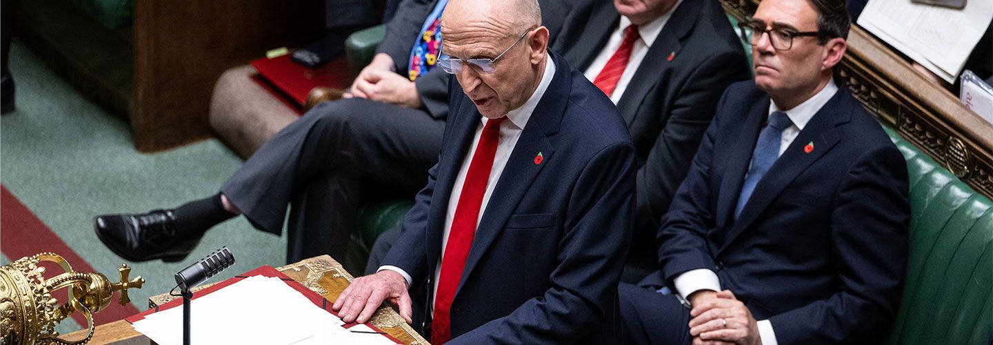 An image of Chancellor Healey in the House of Parliament delivering the Budget. He wears a smart dark blue suit and red tie and small poppy pin on his suit. Sitting behind him can be seen PM Burnham. In front of him is the corner of a crown