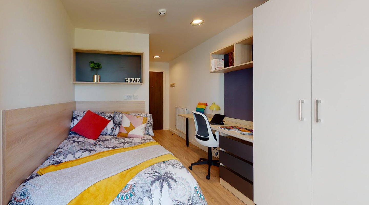 Single bed, wardrobe and desk. Pale laminate floor, cream walls and furniture in pale wood and black. Radiator and coat hooks by the door at the far end.