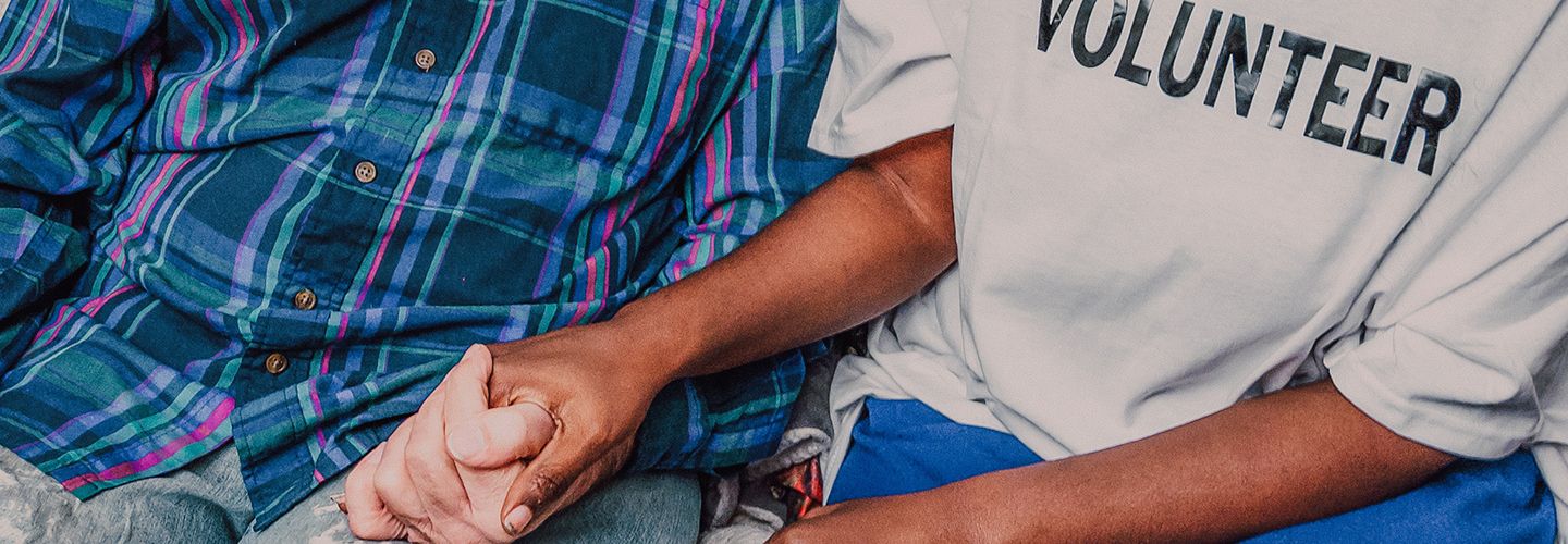 Woman wearing a top with 'VOLUNTEER' written across the chest sits on a sofa with a man and holds his hand.