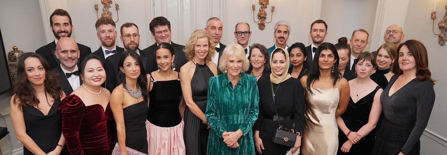Queen Camilla stands at the centre of a group of award-winning journalists in a green dress. Around her stand journalists in black tie attire.